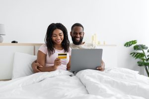 Happy young black woman and husband with computer and credit card sitting on bed in bedroom interior